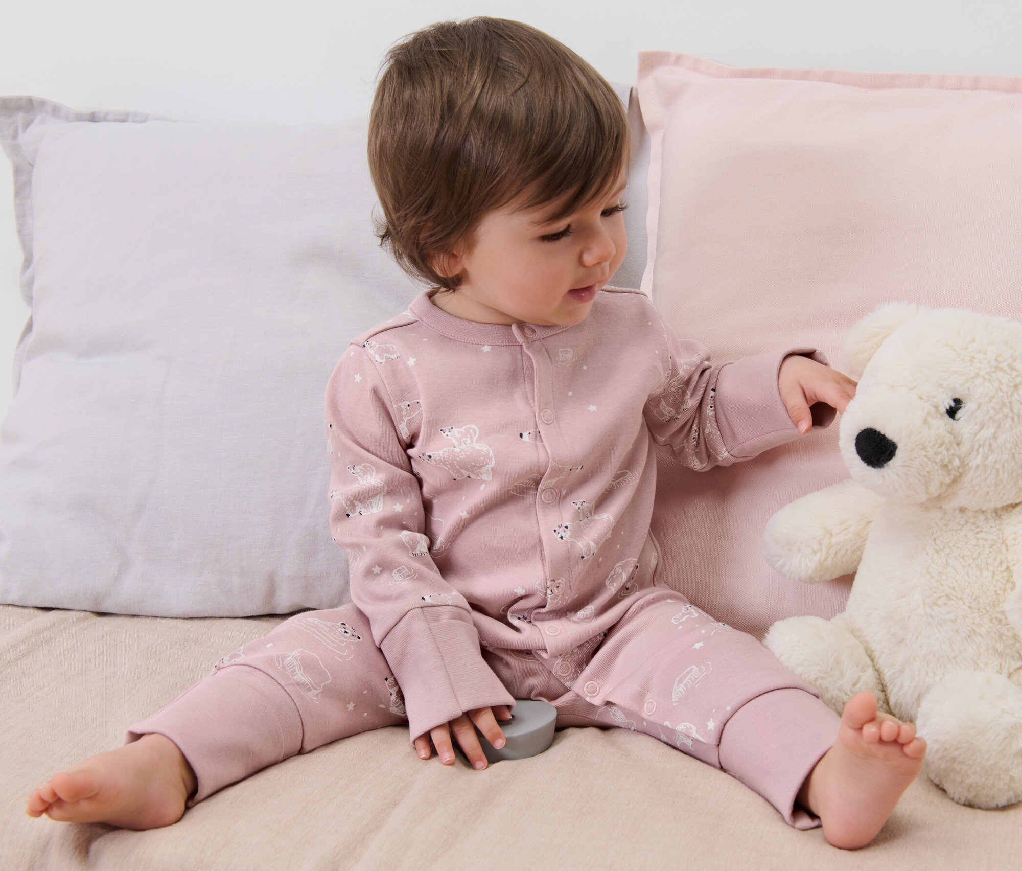 Ein Kind sitzt auf einem Bett und spielt mit einem Teddybär. Es trägt einen rosa 3er-Pack Baby-Pyjamas.