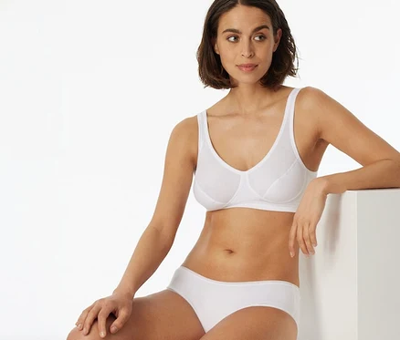 Eine Frau mit kurzen dunklen Haaren trägt ein weißes SCHIESSER Bustier mit Cups und einen weißen Slip. Sie sitzt auf einem weißen Block und lehnt sich an einen anderen weißen Block.
