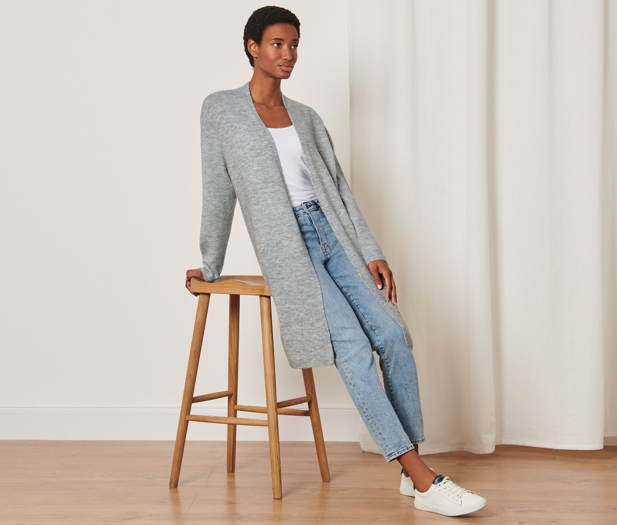 Frau sitzt auf einem Hocker und trägt einen offenen Longcardigan mit RWS-Wolle, Jeans und Turnschuhe.