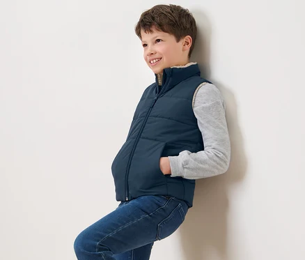 Ein Junge lehnt an einer Wand und trägt eine marineblaue Kinder-Teddyplüsch-Steppweste.