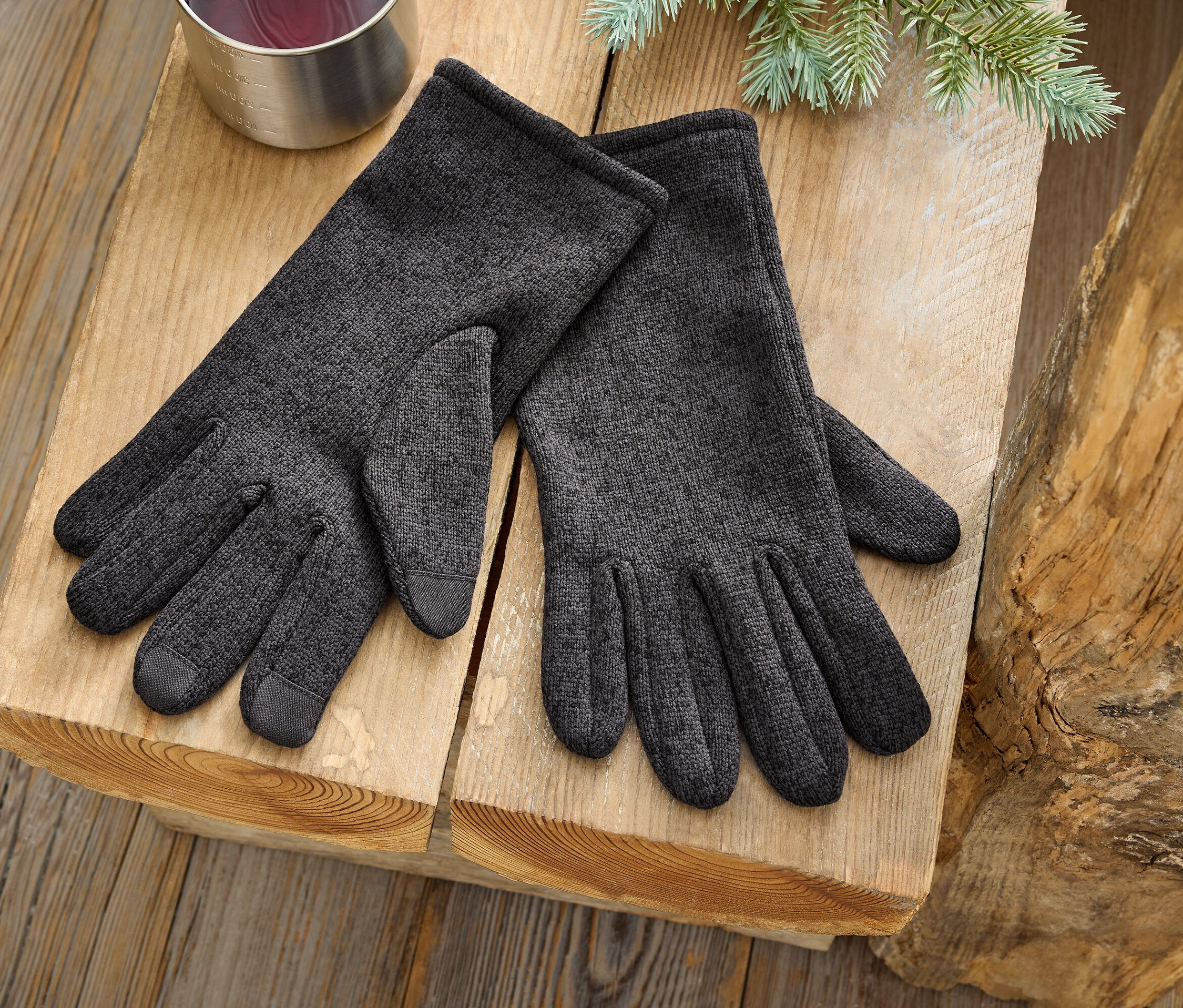 Ein Paar graue Handschuhe liegen auf einer Holzoberfläche neben einem silbernen Becher und einem Nadelbaumzweig.