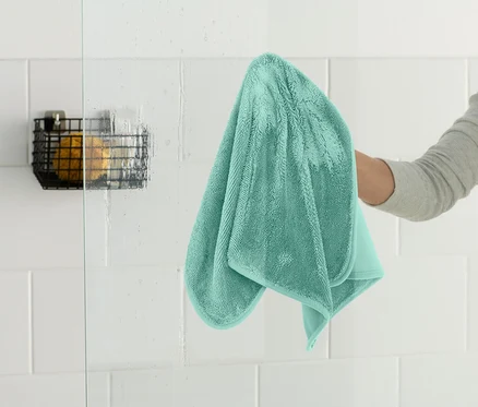 Eine Person wischt eine gläserne Duschwand mit einem türkisfarbenen Handtuch ab. In der Ecke befindet sich ein schwarzer Drahtkorb.