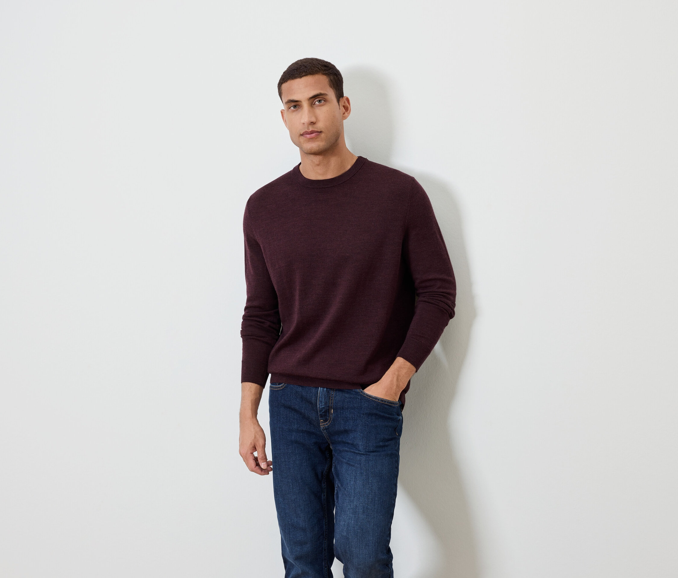 Mann posiert vor einem weißen Hintergrund und trägt einen burgunderfarbenen Pullover und Jeans.