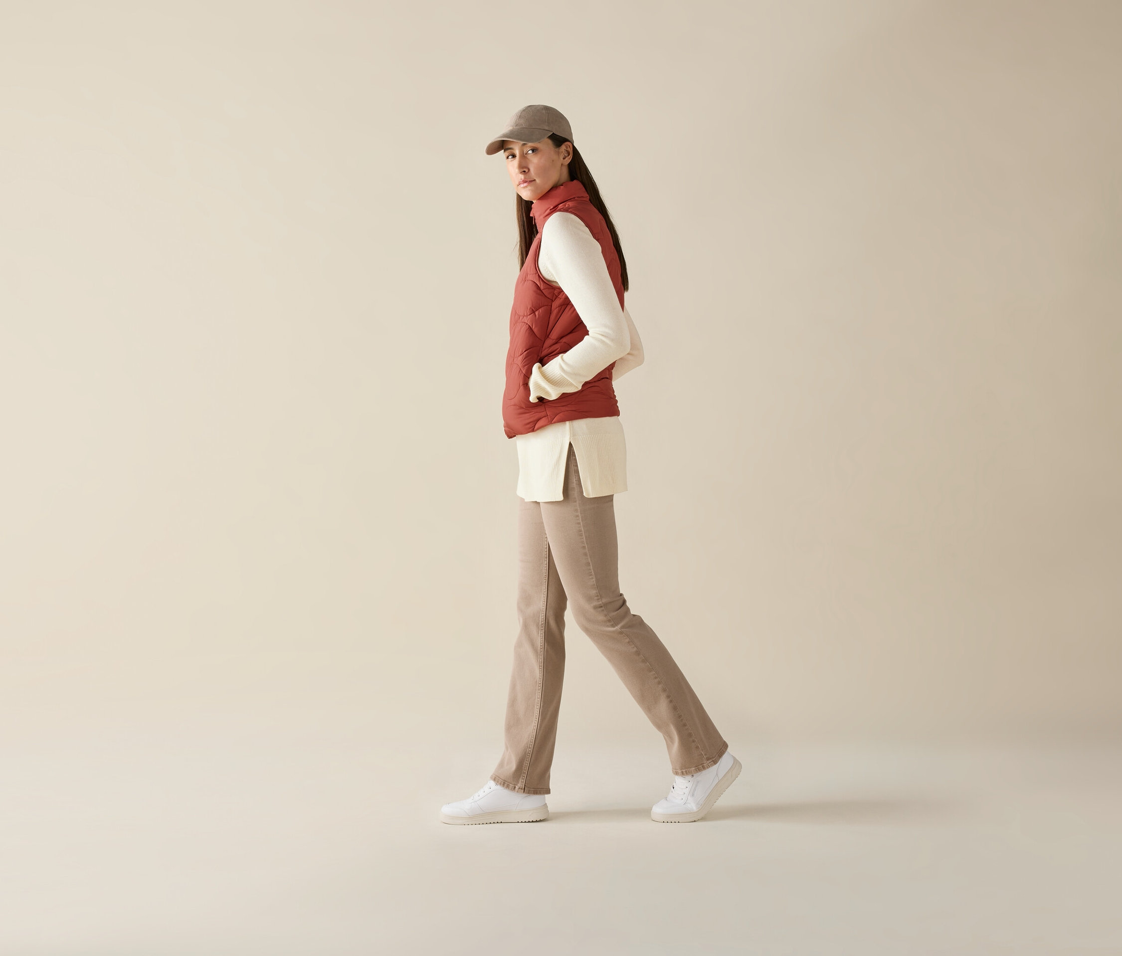 Frau geht, trägt eine Steppweste, ein langärmliges Oberteil, Hosen und eine Schirmmütze.