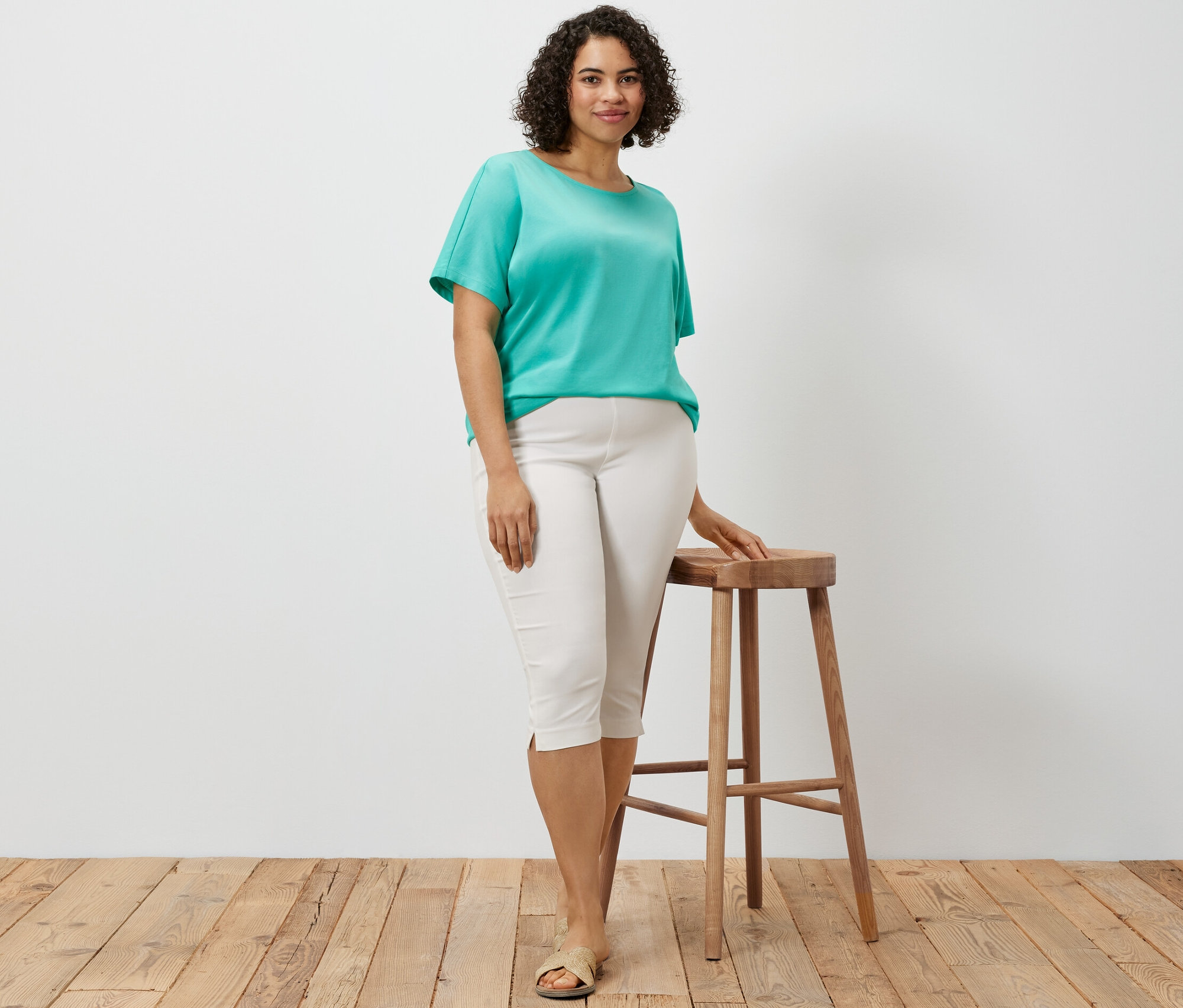 Frau posiert an einem Holzschemel, trägt eine weiße 3/4-Stretchhose und ein türkisfarbenes, überschnittenes Blusenshirt.