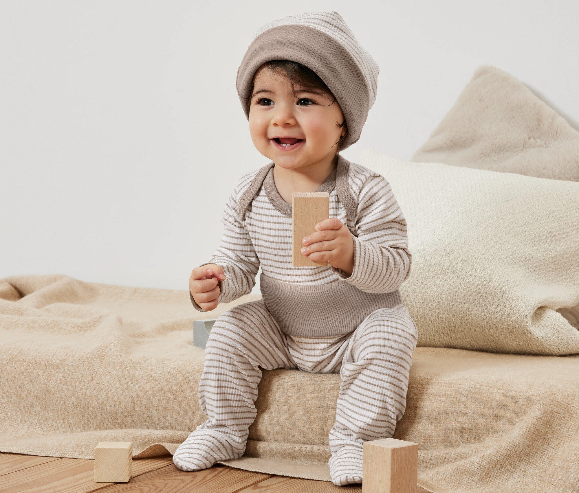 Ein lächelndes Baby sitzt auf einer Decke und spielt mit Holzklötzen. Es trägt ein gestreiftes Baby-Set, 3-teilig.