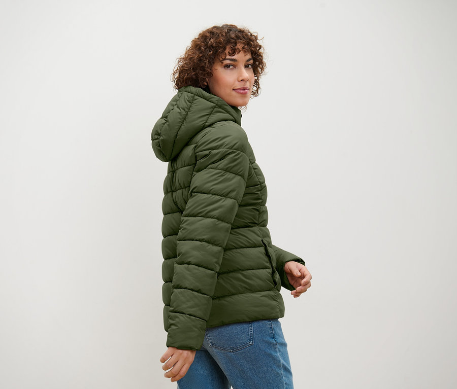 Frau mit lockigem Haar schaut über die Schulter und posiert in einer olivgrünen Steppjacke mit Kapuze und Jeansjacke.