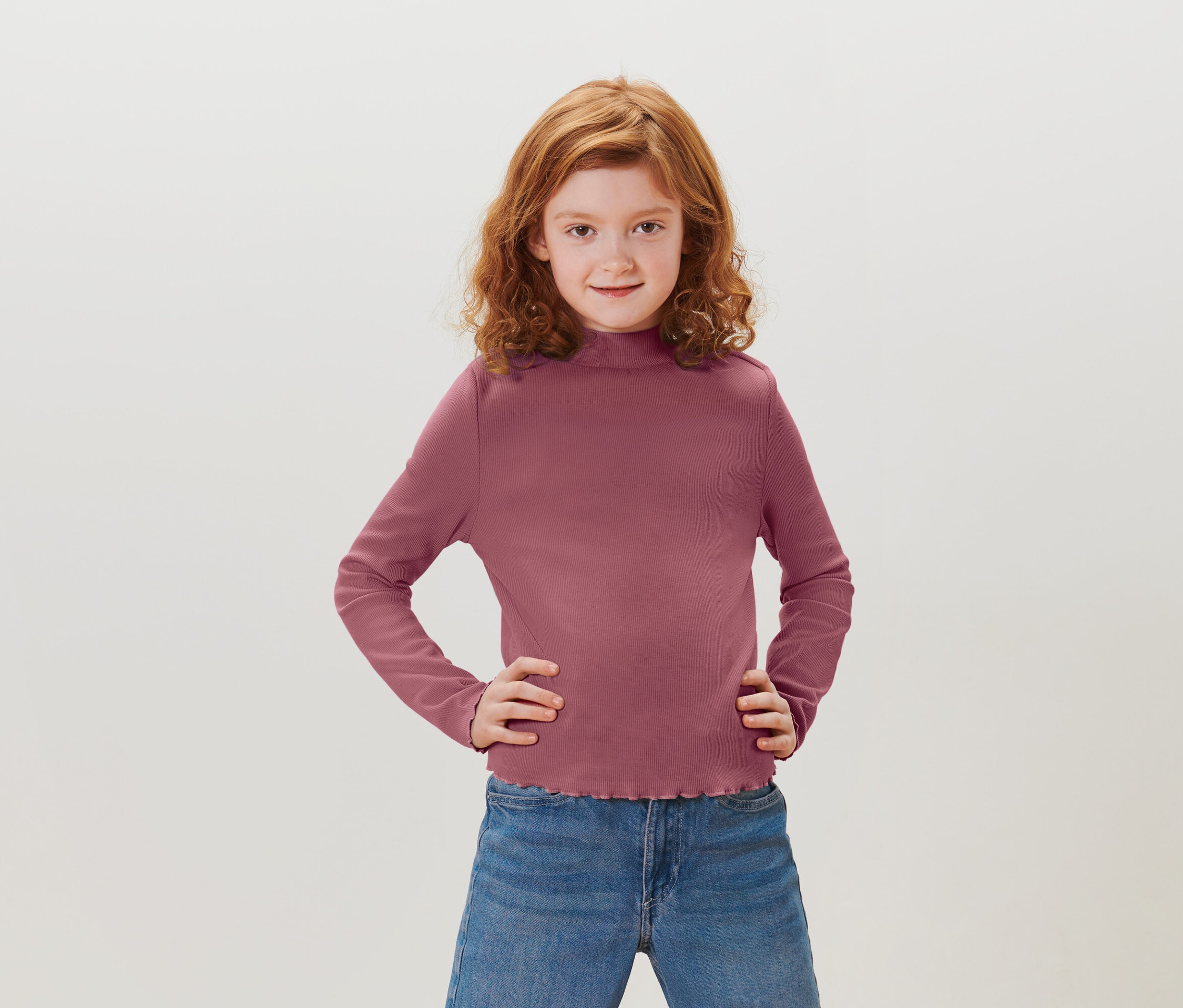 Ein Mädchen mit roten lockigen Haaren steht mit den Händen in den Hüften. Sie trägt ein purpurrotes Langarmshirt und blaue Jeans.