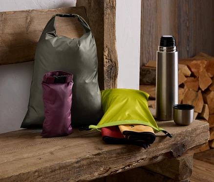 Auf einer Holzbank liegen ein grüner Sack, ein lila Sack, ein grüner Sack mit Tüchern, eine Thermoskanne und ein Holzstapel.