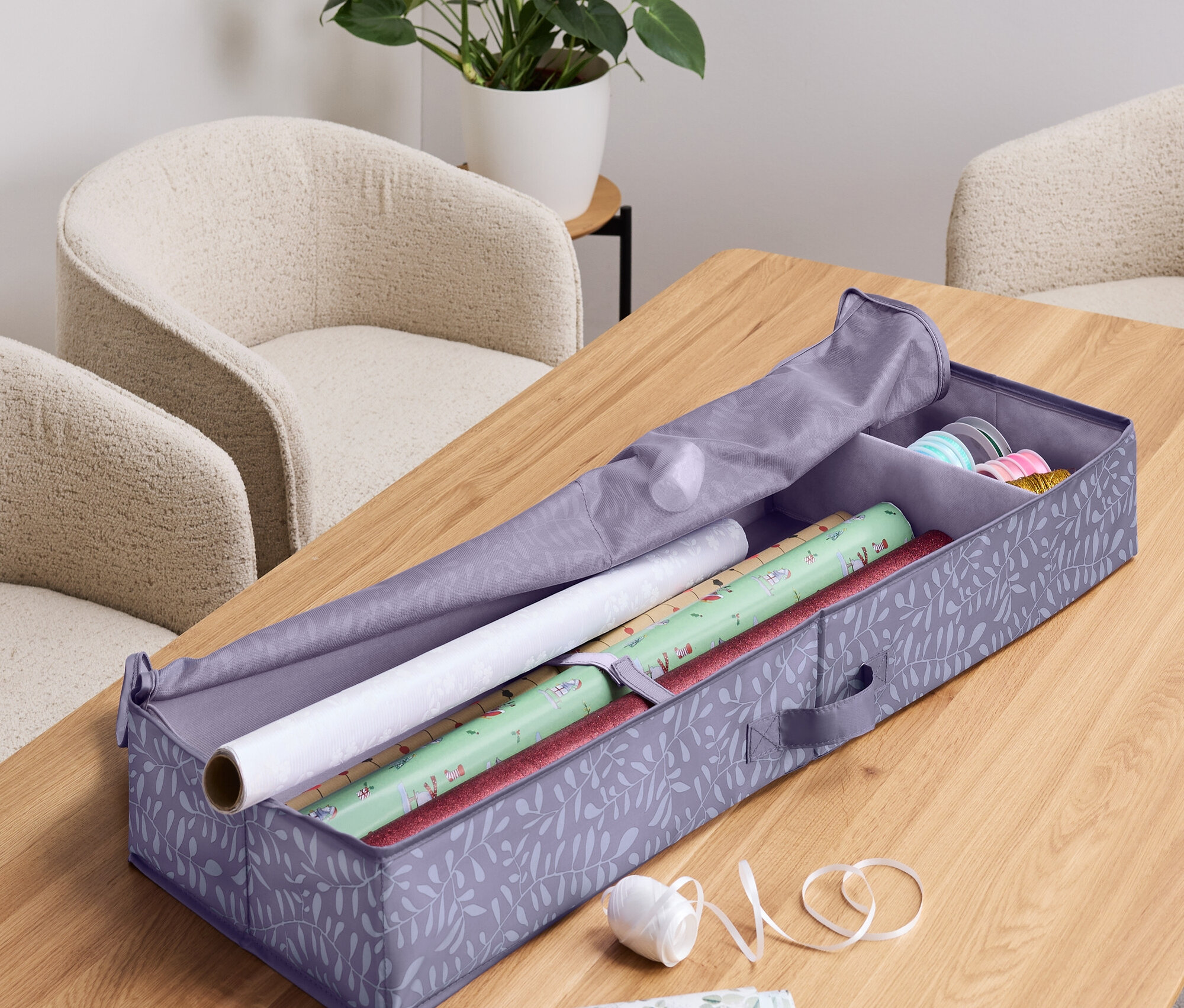 Offene Aufbewahrungsbox für Geschenkpapier mit Papierrollen, Band, Schere und Pflanze im Hintergrund.