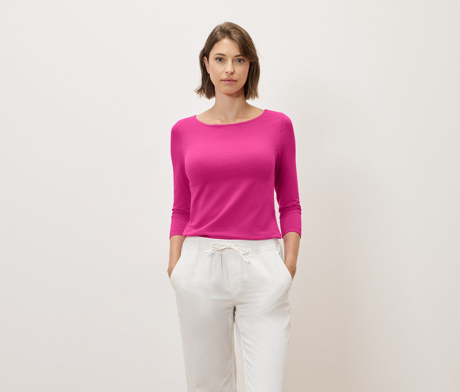 Frau mit braunen Haaren posiert in einem pinkfarbenen Oberteil mit Dreiviertelärmeln und weißen Hosen.