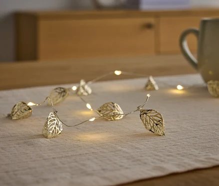 Eine goldene Lichterkette in Blattform liegt auf einem Tischläufer neben einer Tasse.