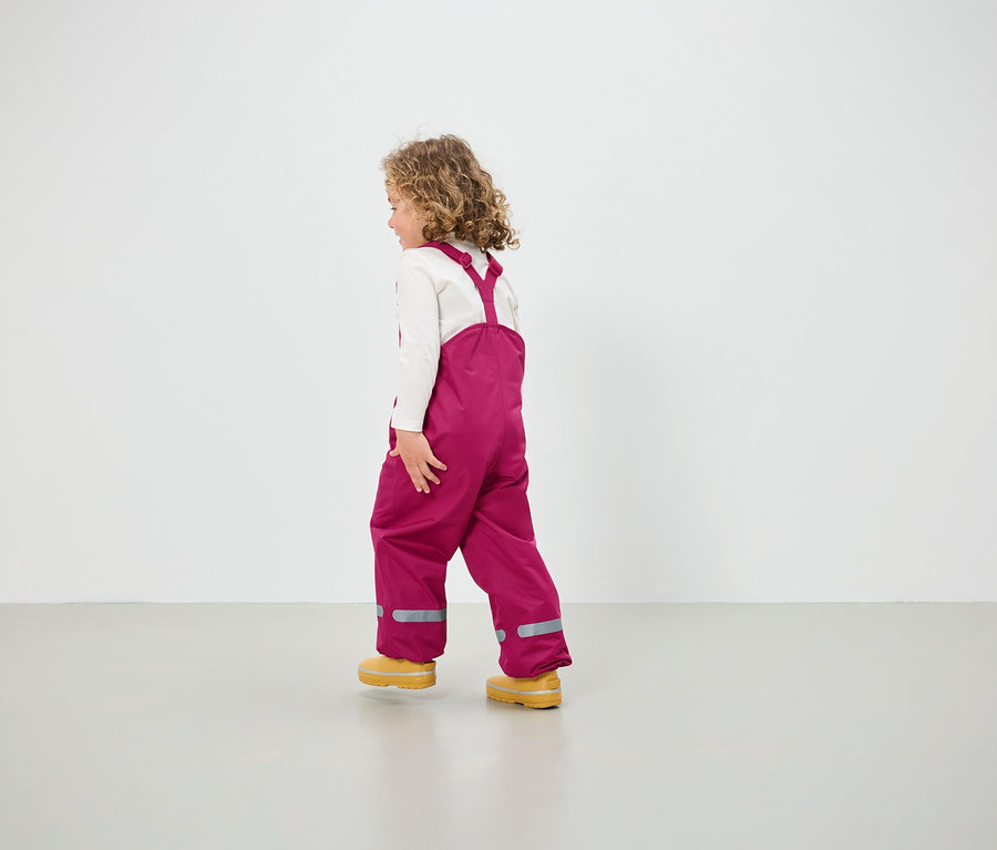 Mädchen in pinkfarbener Latzhose und gelben Schuhen geht über den Boden.