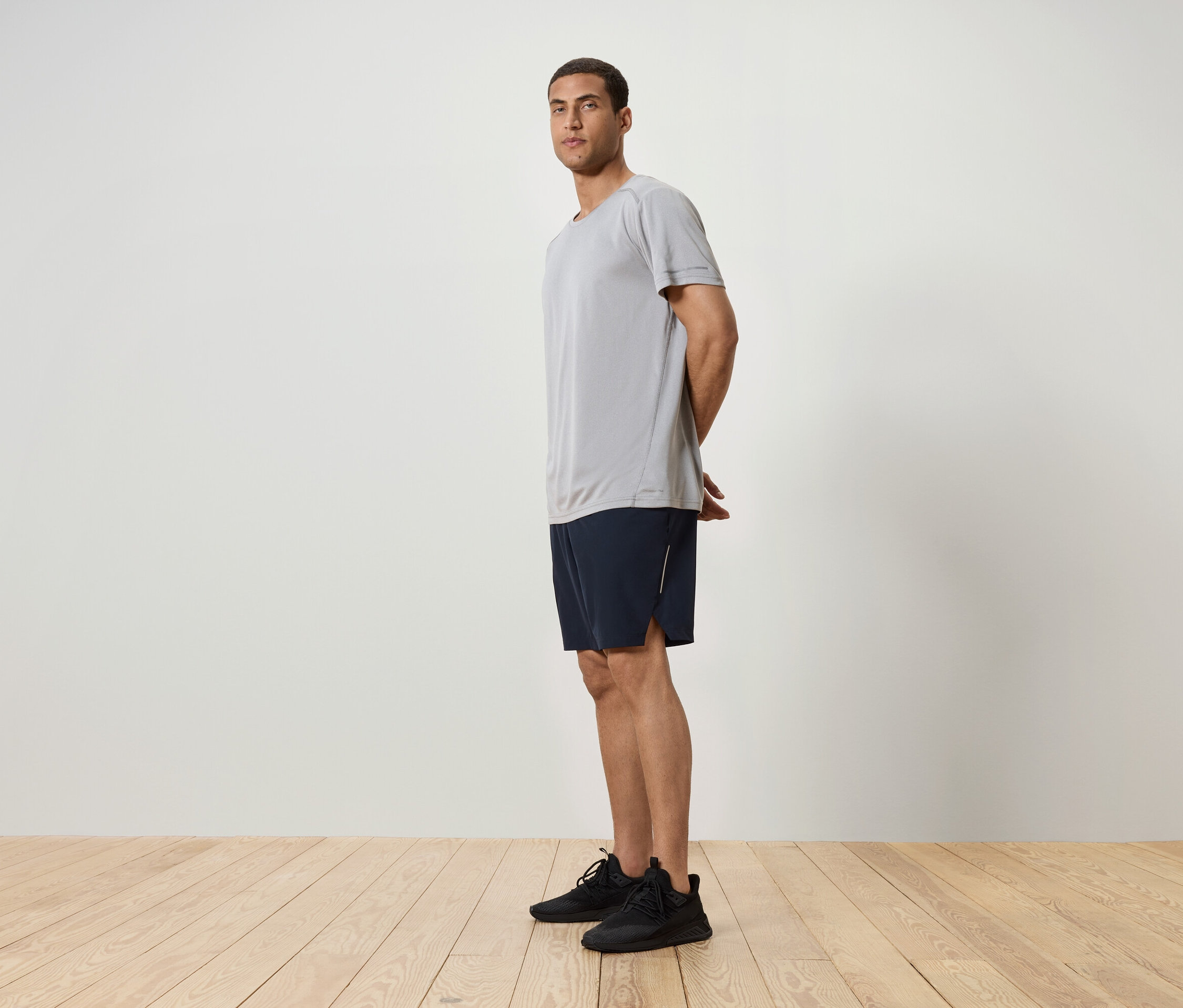 Mann steht seitlich, trägt ein graues T-Shirt, dunkelblaue Shorts und schwarze Turnschuhe.