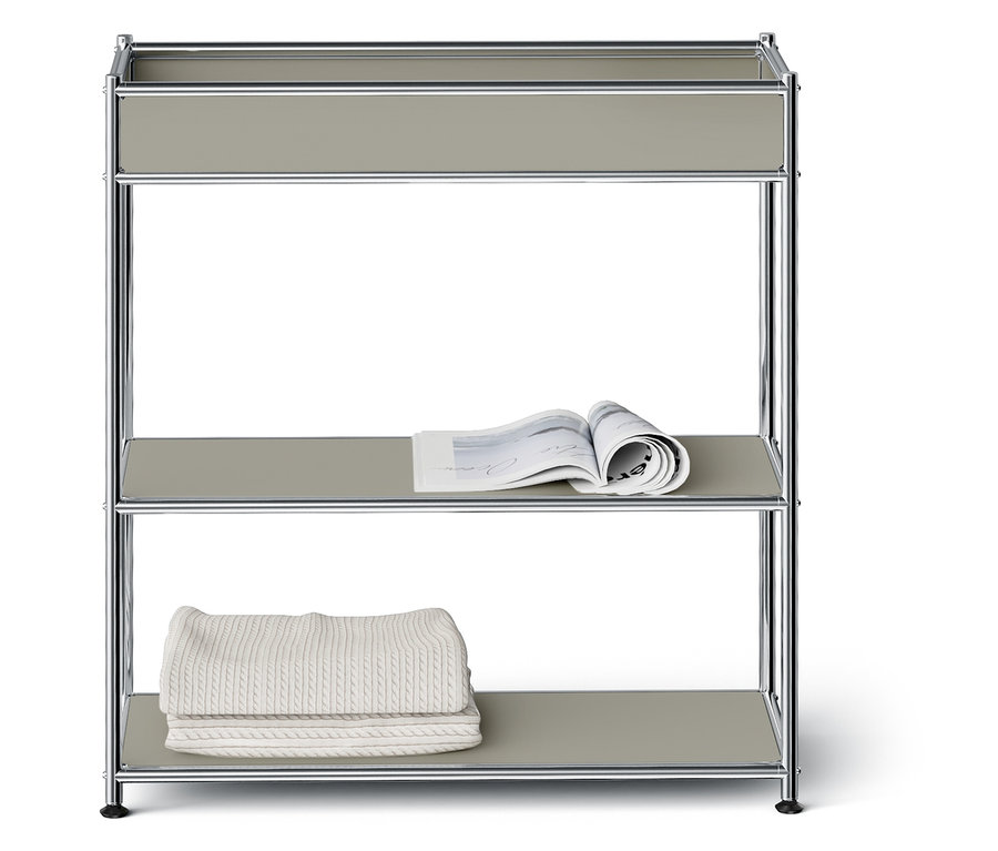 Ein dreistöckiges Regal mit silbernem Rahmen und grauen Regalböden. Auf dem mittleren Regal liegt eine offene Zeitschrift, auf dem unteren Regal liegt eine gefaltete weiße Decke.