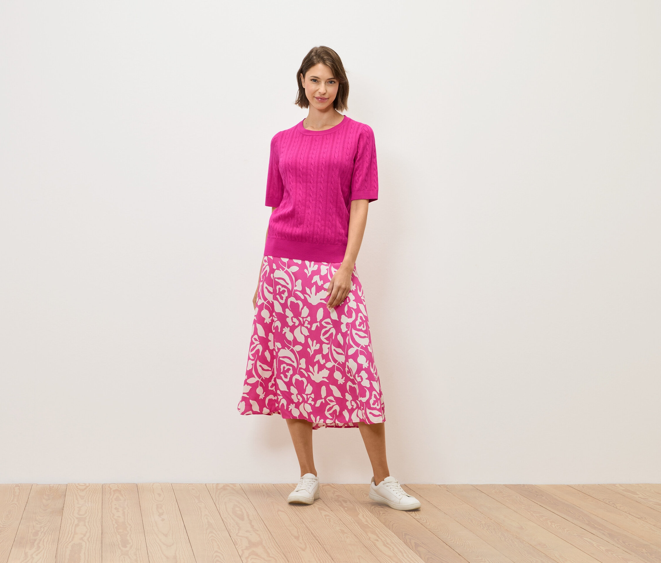 Eine Frau in einem rosa Pullover und einem Rock mit Blumenmuster steht auf einem Holzboden.