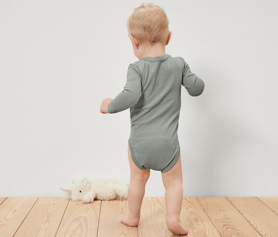 Kleines Kind in einem gerippten Langarmbody steht auf einem Holzboden neben einem Plüschtier.