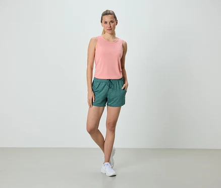 Frau steht mit den Händen in den Taschen in grünen Shorts und einem rosa Tanktop.