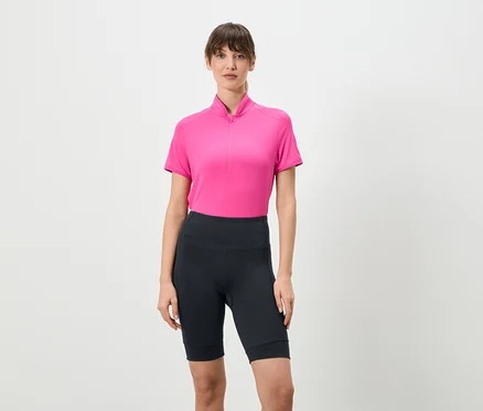 Frau steht und trägt ein pinkes Shirt mit kurzem Reißverschluss und kurze, dunkle Leggings vor einem weißen Hintergrund.