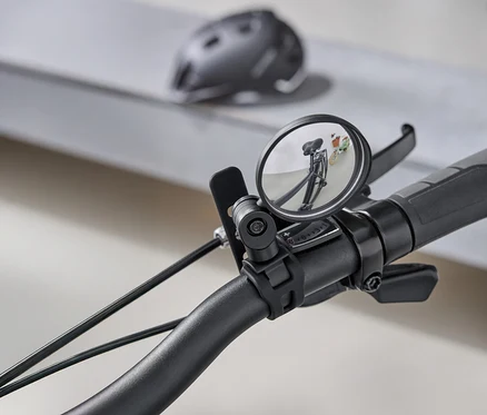 Nahaufnahme eines Fahrradlenkers mit einem runden Rückspiegel, der den dahinterliegenden Weg reflektiert. Im Hintergrund ist ein grauer Fahrradhelm zu sehen.
