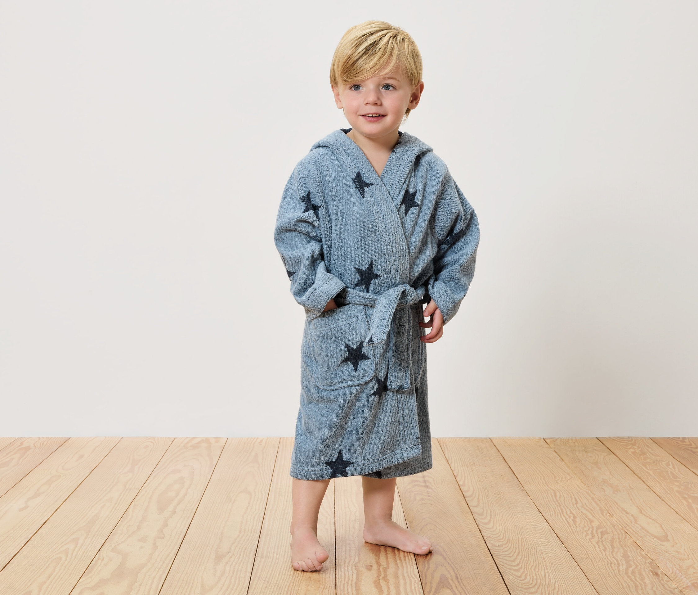Ein kleiner Junge mit blonden Haaren steht barfuß auf einem Holzboden und trägt einen hellblauen Bademantel mit dunklen Sternen.