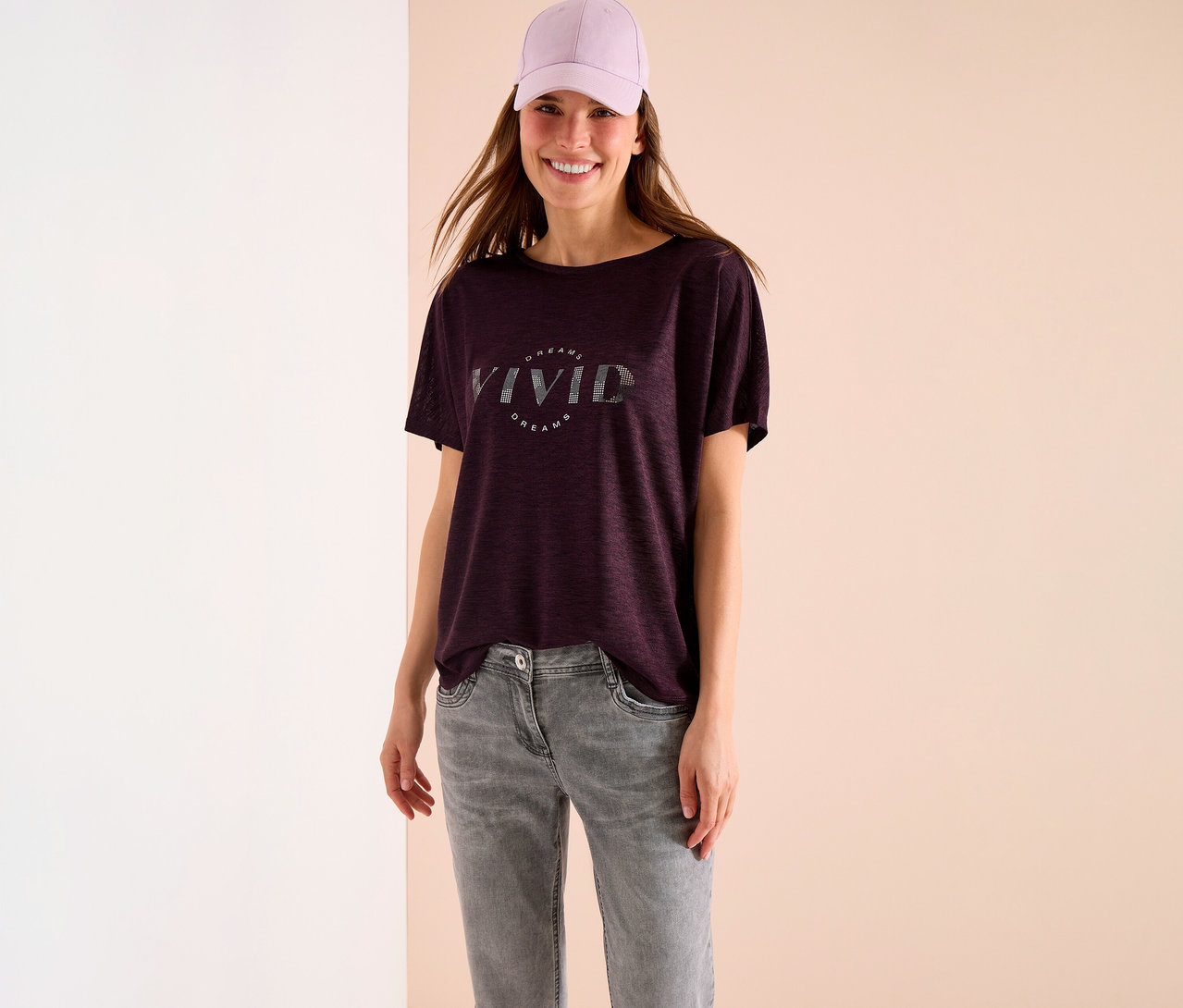Frau mit lila Kappe, dunkelrotem T-Shirt mit "Vivid Dreams" Aufdruck und grauer Jeans steht vor einer Wand.