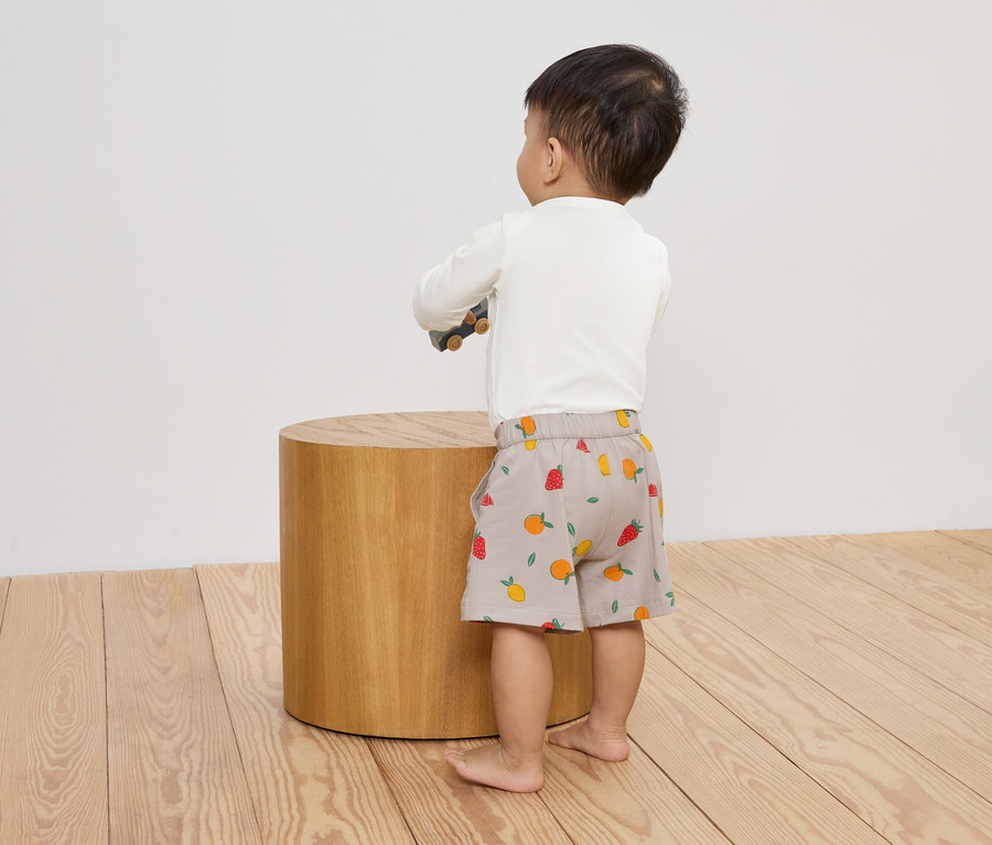 Ein Kleinkind steht mit dem Rücken zum Betrachter auf einem Holzboden und stützt sich mit einem Spielzeugauto auf einem runden Holzschemel ab.