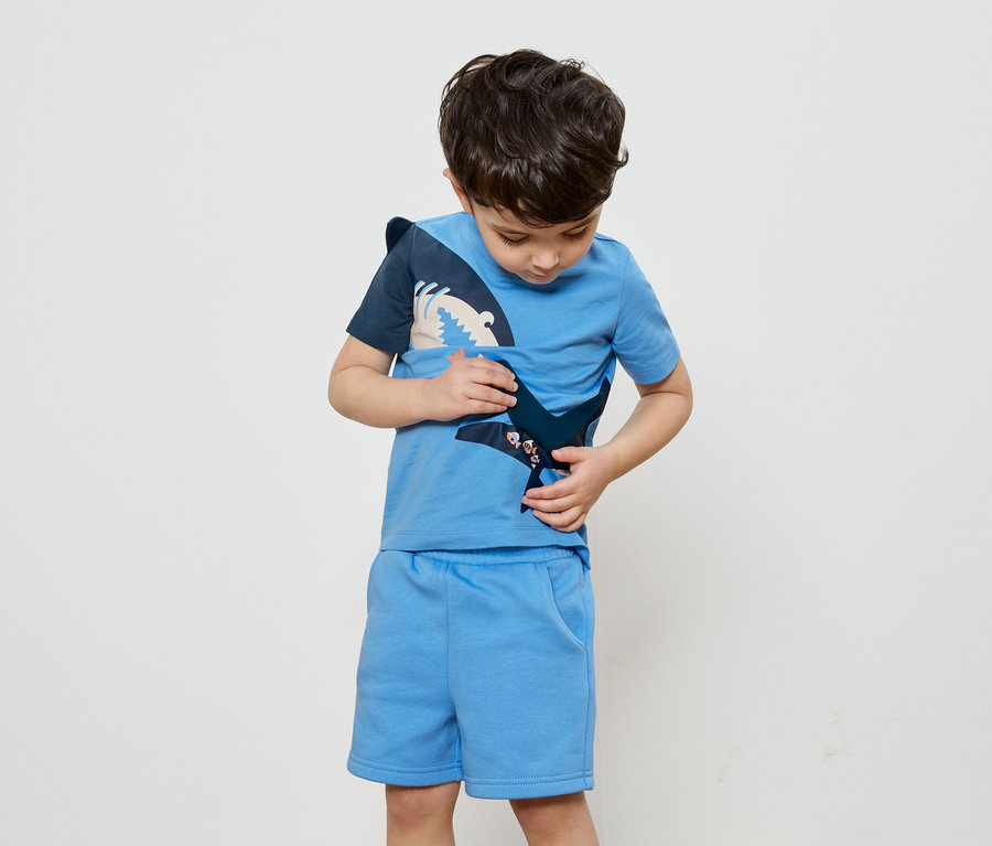 Ein Junge betrachtet ein blaues T-Shirt mit Hai-Applikation, dazu passende Shorts.