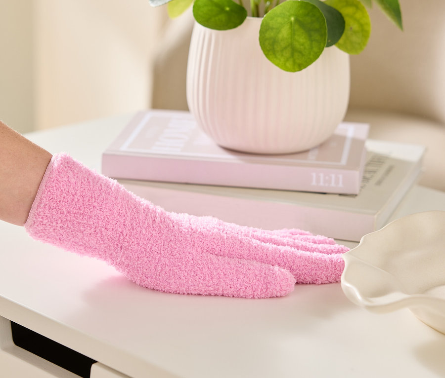 Eine Hand mit einem rosa Chenille-Handschuh wischt über eine weiße Oberfläche, im Hintergrund eine Zimmerpflanze und Bücher.