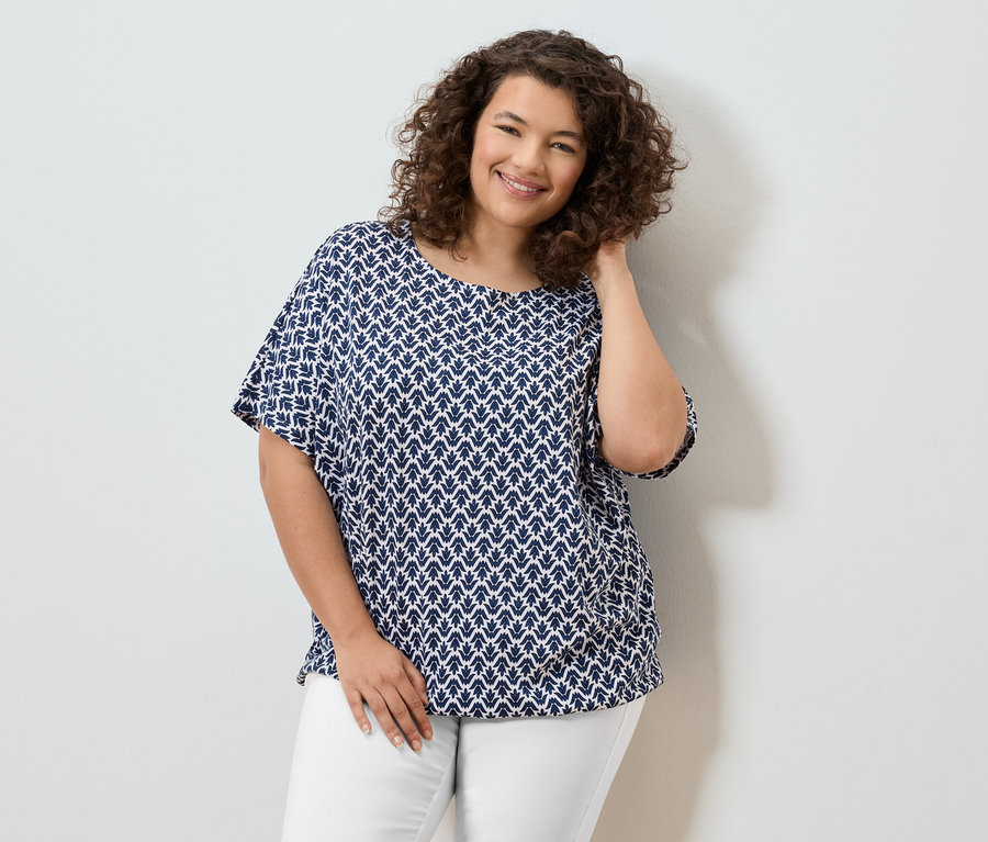 Frau mit lockigem Haar, die eine blau-weiße Bluse und weiße Hosen trägt und lächelt.