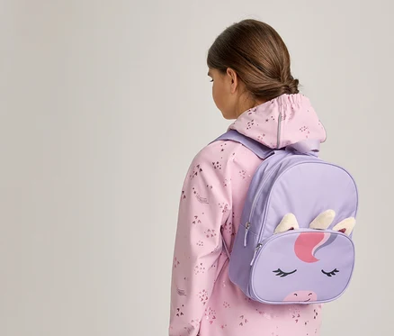 Ein junger Mensch von hinten gesehen, trägt eine rosa gemusterte Jacke und einen lila Einhorn-Rucksack mit Einhorn-Gesicht, Ohren und Horn.
