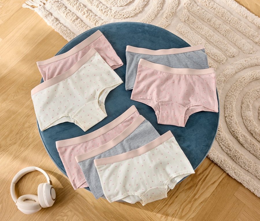 Auf einem blauen Hocker liegen sechs Damenshorts in Grau, Rosa und mit Punktmuster, daneben Kopfhörer.