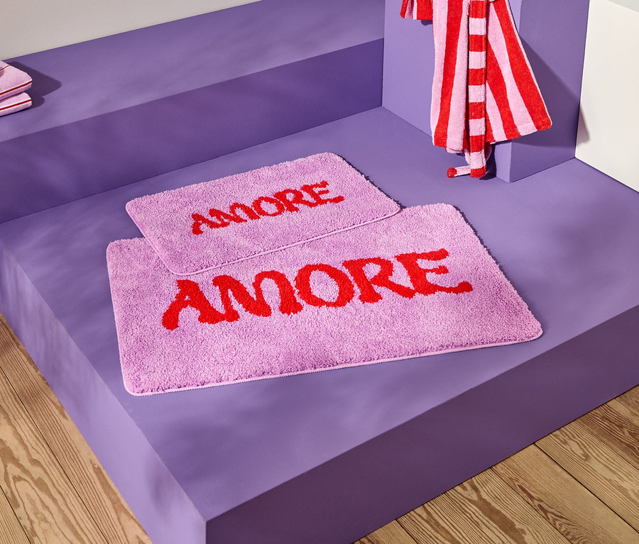 Aufnahme von zwei lavendelfarbenen Badematten mit dem roten Wort "AMORE" auf einer violetten Oberfläche, zusammen mit gestreiften Handtüchern.