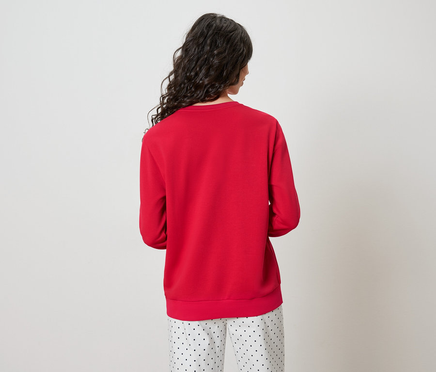 Rückansicht einer Frau mit langen, gewellten, dunklen Haaren, die einen roten Pullover und eine gepunktete Hose trägt.