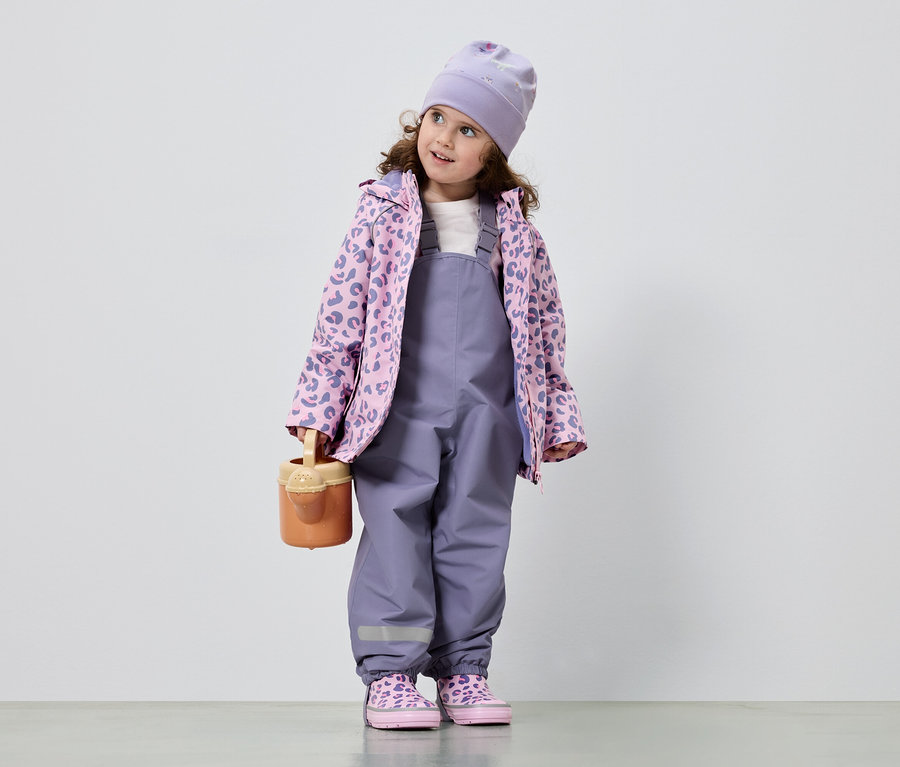 Ein kleines Mädchen mit lockigem Haar trägt einen lila Overall, eine rosa-lila Jacke mit Leopardenmuster, eine Mütze und passende Schuhe. Sie hält eine Gießkanne.
