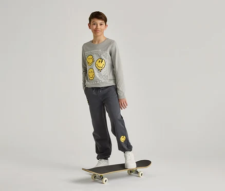 Ein Junge mit grauem Sweatshirt und Hose mit gelben Smileys steht auf einem Skateboard vor einem weißen Hintergrund.