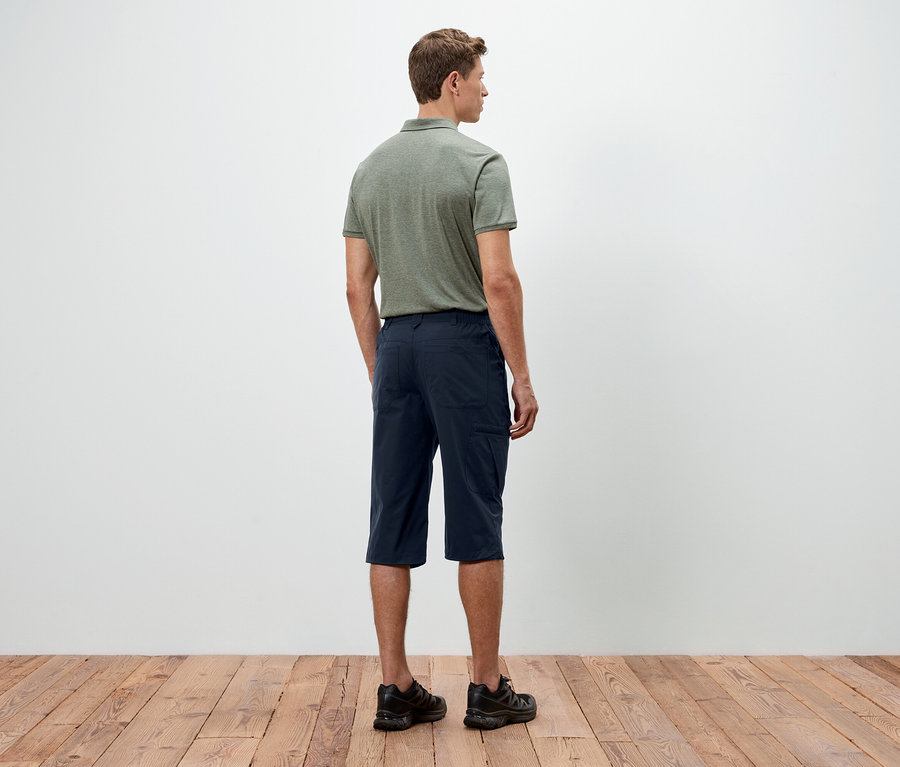 Ein Mann steht mit dem Rücken zur Kamera, er trägt ein grünes Polohemd, blaue Shorts und schwarze Schuhe.