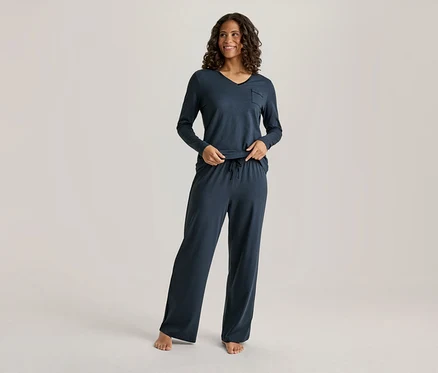 Eine lächelnde Frau mit dunklem, lockigem Haar trägt ein dunkelblaues Langarm-V-Neck-Oberteil mit Tasche und eine passende weite Hose mit Kordelzug. Ihre Hände sind an ihrer Taille, und sie ist barfuß.