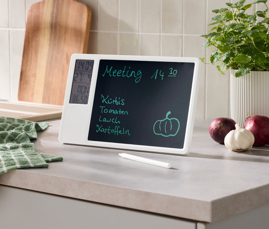 Weißes digitales Memoboard auf einer Küchenarbeitsplatte zeigt Meeting, Einkaufsliste, Uhrzeit, Datum und Temperatur. Daneben liegen Stift, grünes Tuch, Holzschneidebrett, Topfpflanze, rote Zwiebeln und Knoblauch.