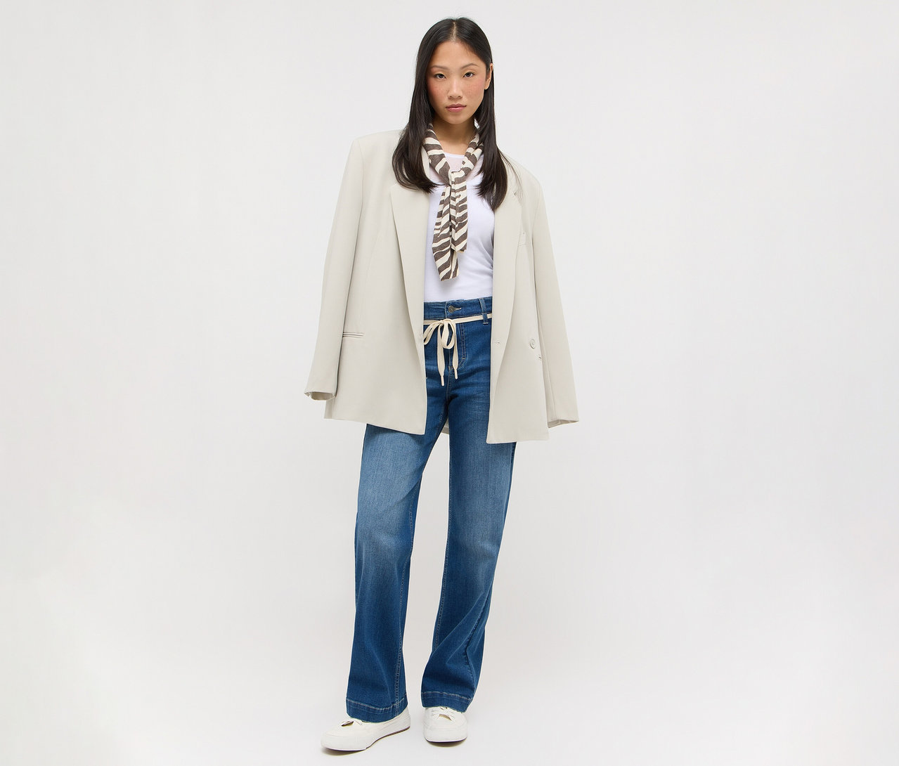 Junge asiatische Frau in weißem T-Shirt, blauen weiten Jeans mit hellem Kordelzug, beigem Oversize-Blazer über den Schultern und braun-weiß gemustertem Schal. Sie trägt weiße flache Schuhe.