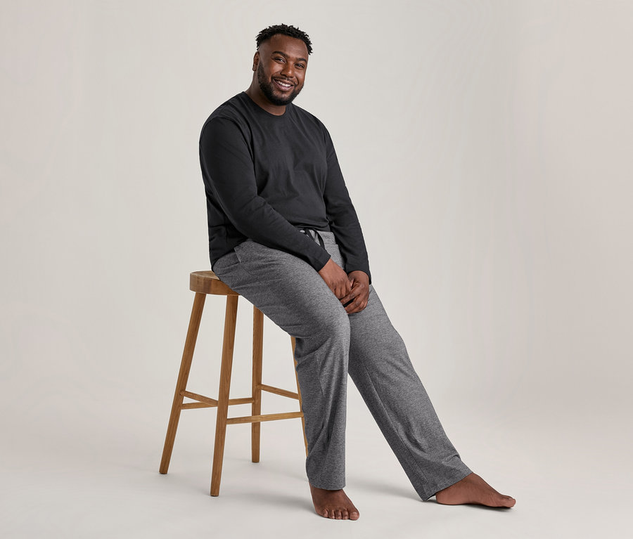 Lächelnder Mann sitzt auf einem Hocker und trägt ein schwarzes Oberteil und eine graue Hose aus einem Pyjama-Set.