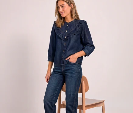 Eine Frau trägt eine dunkelblaue Street One Jeansbluse mit Rüschen und dunkle Jeans, steht neben einem Holzstuhl und blickt zur Seite.