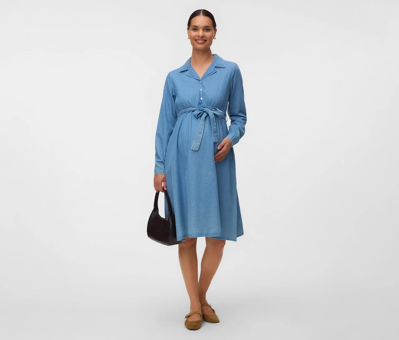 Schwangere Frau in einem blauen Jeanskleid mit Bindegürtel, einer dunkelbraunen Handtasche und hellbraunen flachen Schuhen.