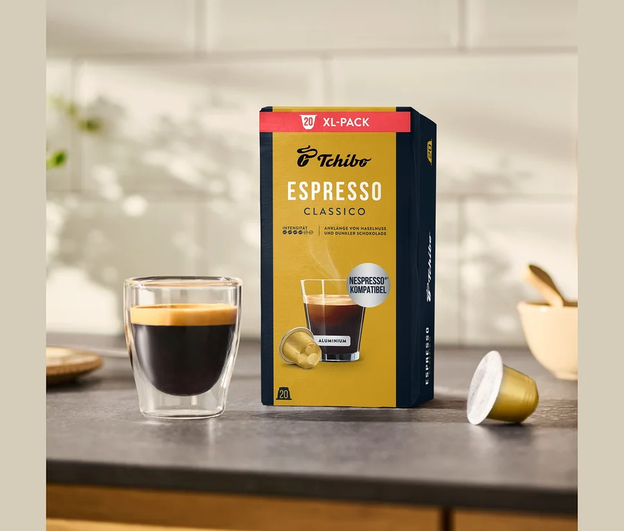 Eine Packung Tchibo Espresso Classico - 20 Kapseln steht neben einem Glas Espresso mit goldener Crema und einer einzelnen Kapsel auf einer Küchenarbeitsplatte.