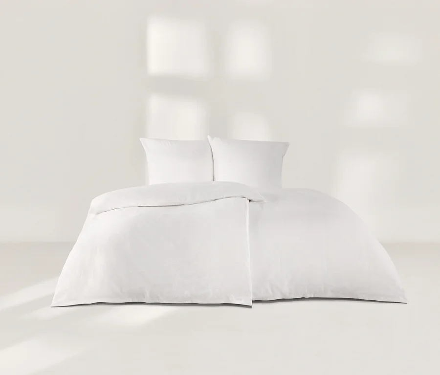 Weisse Leinen-Bettwäsche mit zwei Kissen und einer Bettdecke auf einem Bett vor einer weissen Wand.
