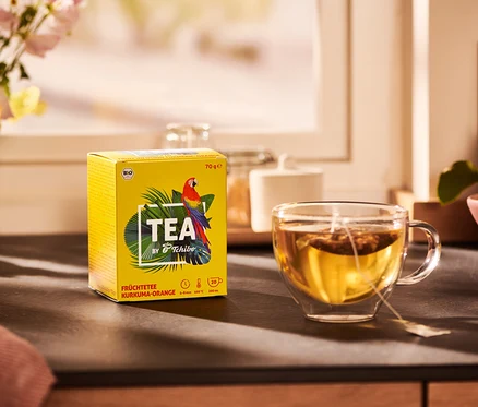 Gelbe Packung TEA by Tchibo Bio Früchtetee Kurkuma-Orange und eine Tasse Tee.