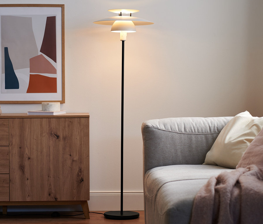 Eine Stehleuchte aus Metall steht neben einer Holzkommode und einem grauen Sofa.
