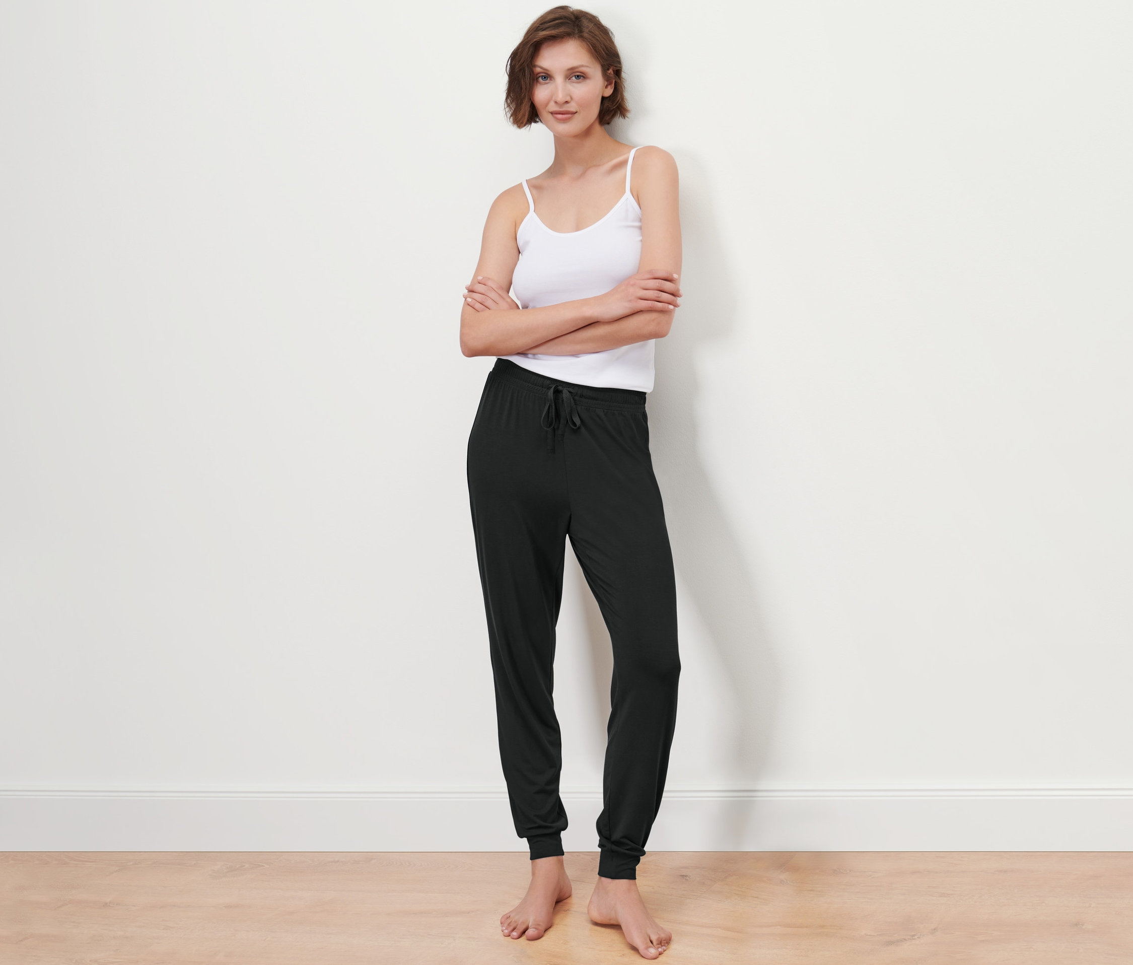 Frau posiert vor einer weißen Wand und trägt ein Tanktop und eine Pyjamahose.