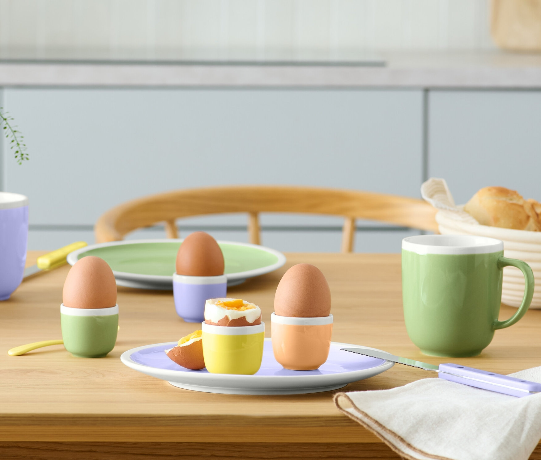 Auf einem Tisch stehen vier Eierbecher mit Eiern, zwei Teller, zwei Tassen, ein Korb mit Brot, eine Serviette und eine Vase mit Blumen.