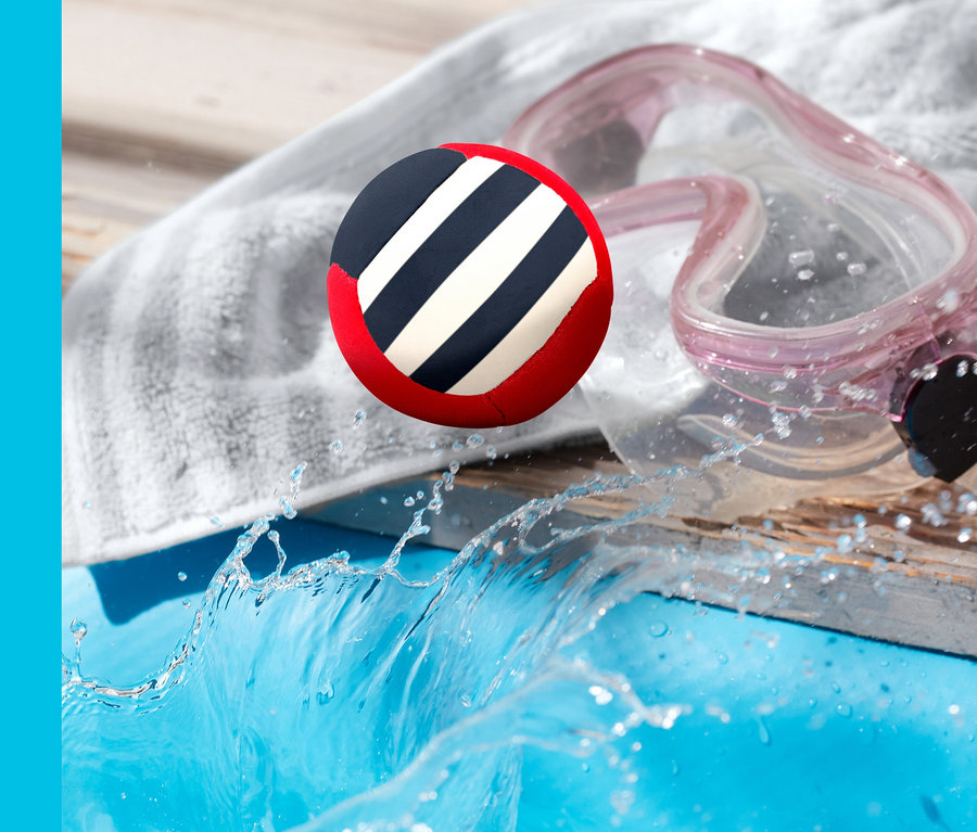 Hüpfender Wasserball spritzt in der Luft über einem Pool, daneben liegen ein Handtuch und eine Schwimmbrille.